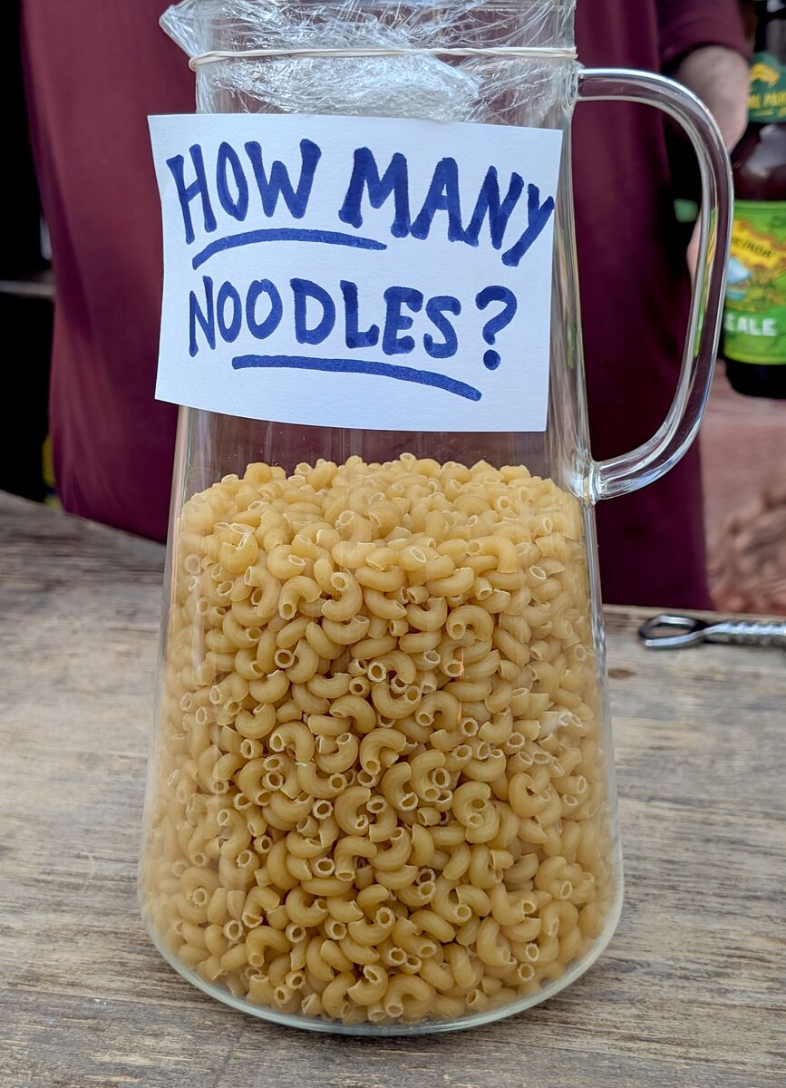 A glass jar filled with elbow macaroni, with a handwritten sign reading 'How many noodles?'